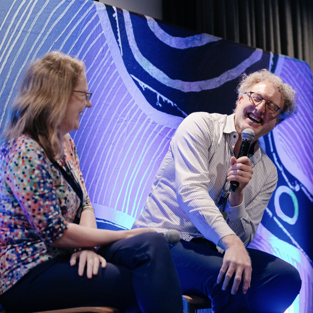 Image of A/Prof. Sally Shrapnel and Prof. Warwick Bowen at SomethingQ in 2024. They are talking on a panel and Warwick is laughing.